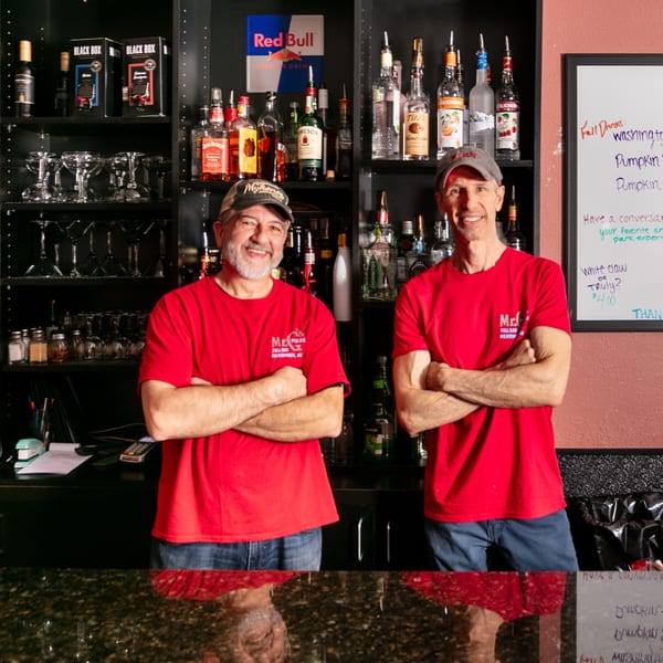 Two smiling men in red Mr. G's Pizzeria shirts stand behind a bar stocked with liquor bottles.