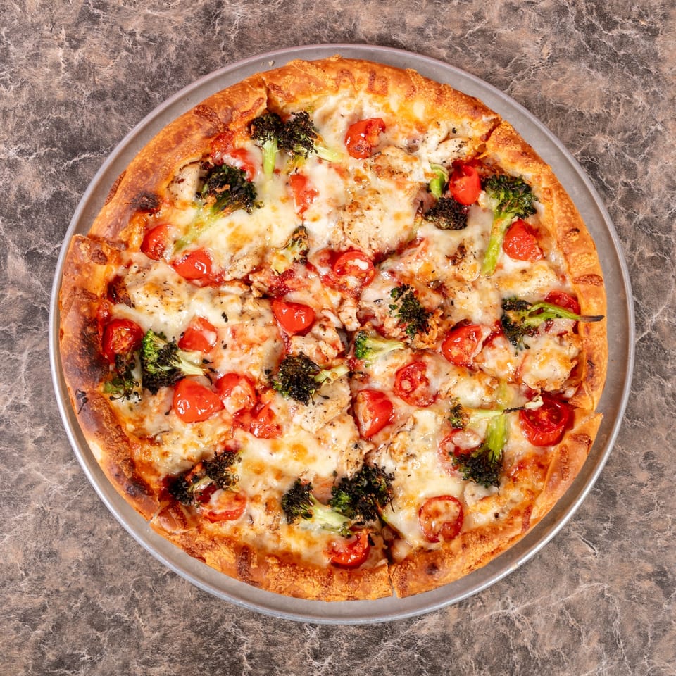 A Mr. G's Pizzeria pizza with melted cheese, cherry tomatoes, and broccoli florets on a metal pan.