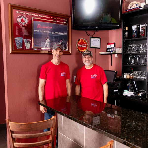 Two smiling Mr. G's Pizzeria staff in red shirts stand behind a dark granite counter.