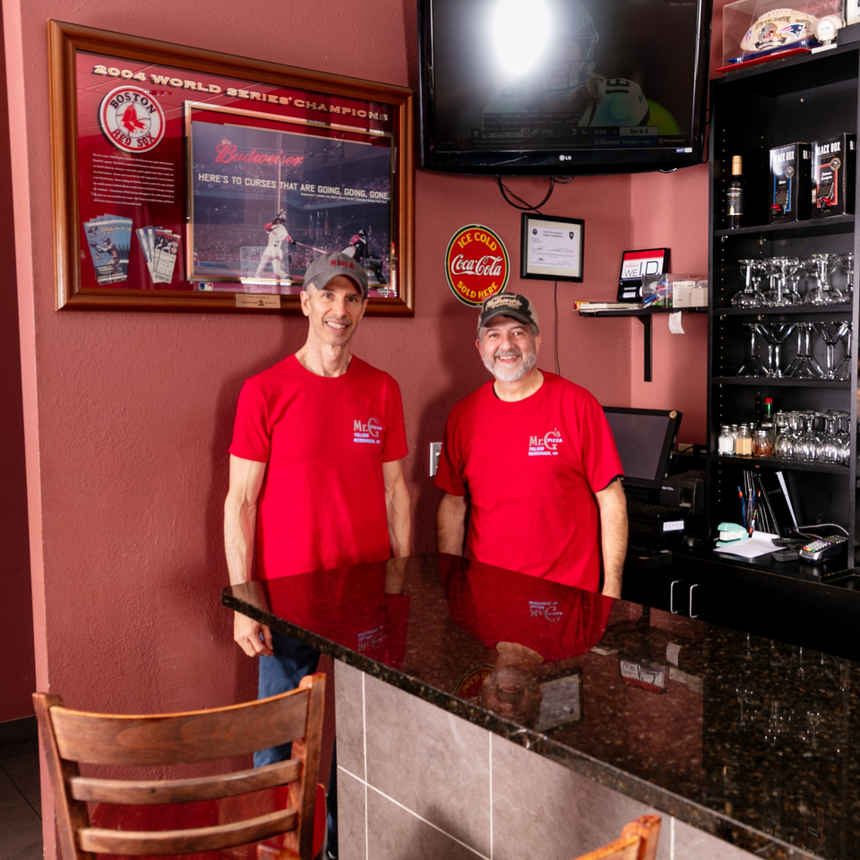 Two smiling Mr. G's Pizzeria staff in red shirts stand behind a dark granite counter.